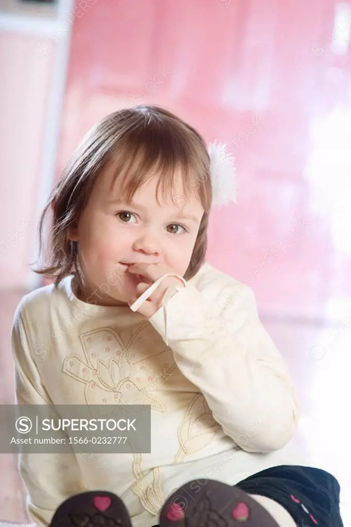 3 year old girl sitting on the floor, smiling into camera, hand to mouth