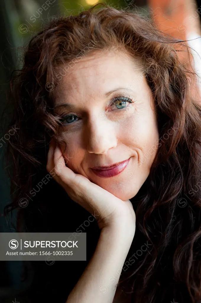 Portrait of a 42 year old woman with long curly black hair smiling at the camera, chin resting in her hand