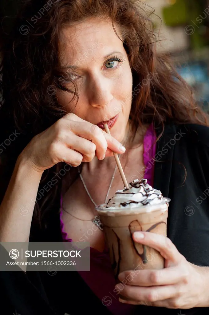 Portrait of a 42 year old woman with long curly black hair sucking on the straw of a cold coffee drink
