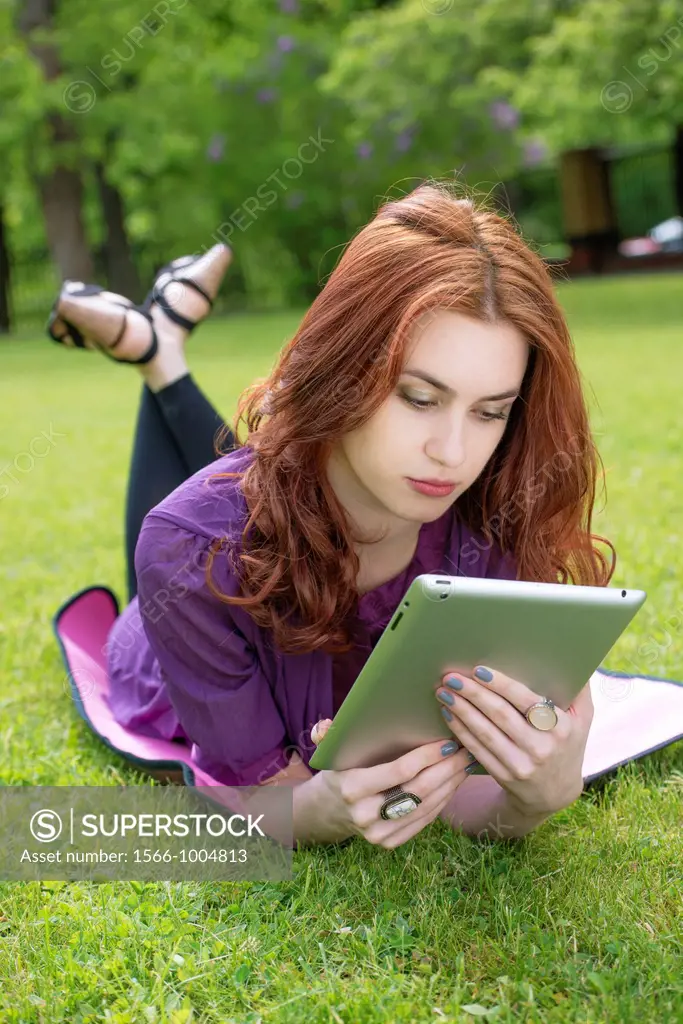 Young woman lying on the grass in a park and reading on a tablet pc
