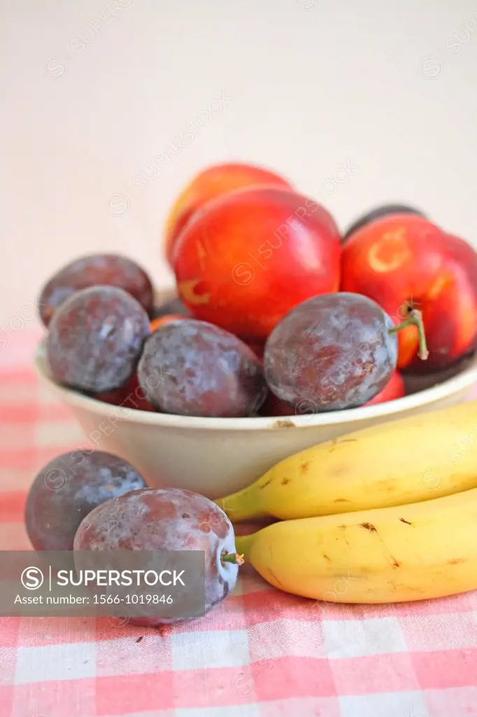 Plums and a small green apple in a white ceramic with bananas  Fresh purple plums in an white bowl  Small green apple windfall  Bananas lay beside the...