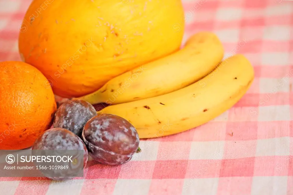Melon, bananas, plums and orange on table  Fruit is laid on red gingham tablecloth in artful manner