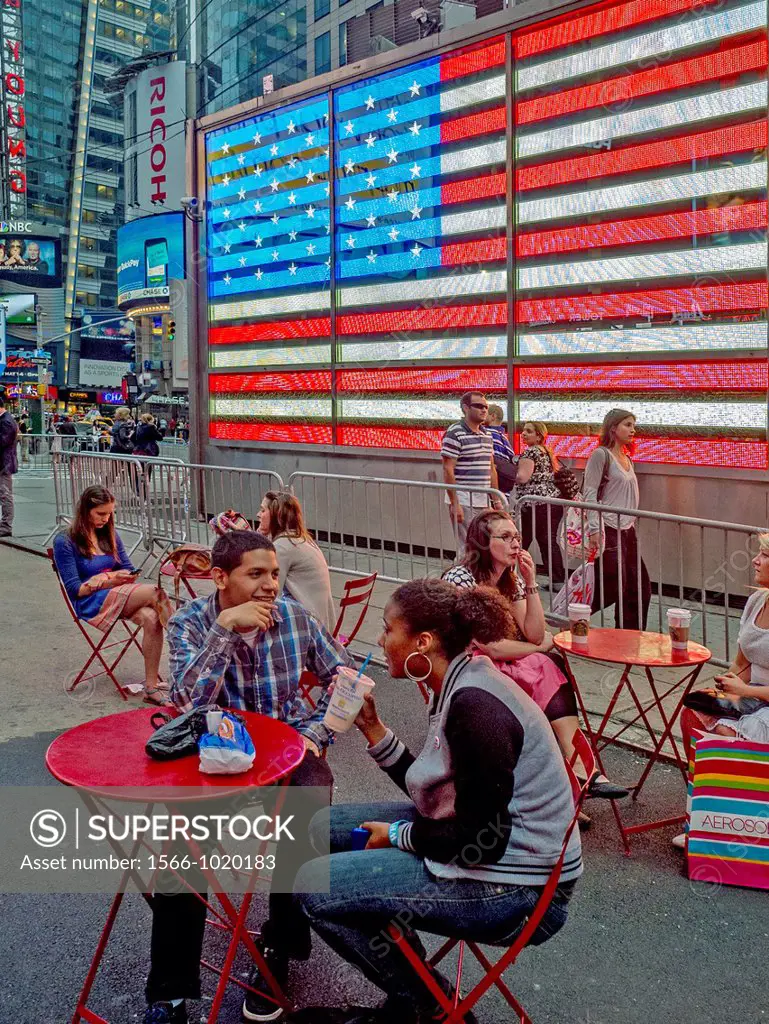 People relax at outdoor tables in Times Square, New York City  In background is the electric United States flag on the wall of a military recruiting c...
