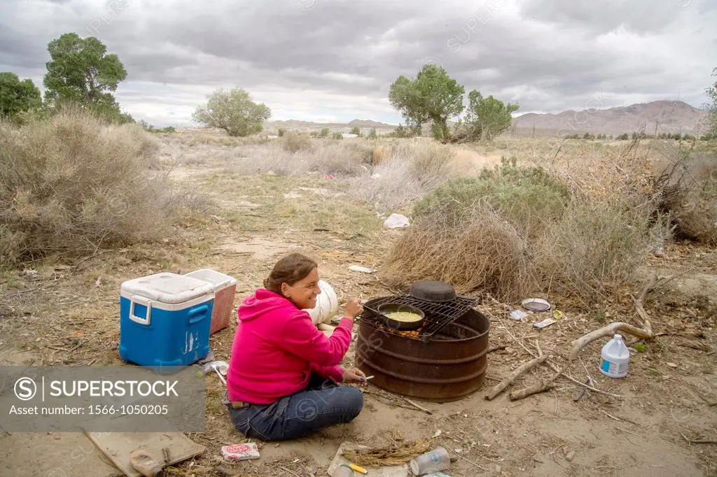 A homeless woman cooks corn on a primitive outdoor grill a primitive outdoor encampment in the desert town of Victorville, CA