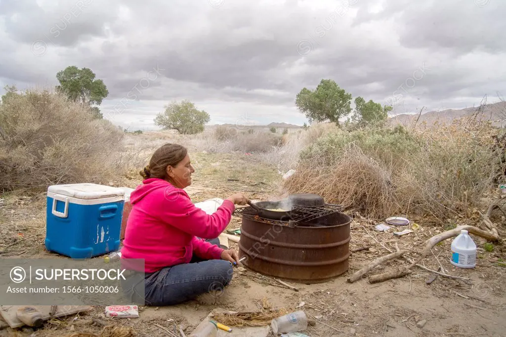 A homeless woman cooks corn on a primitive outdoor grill a primitive outdoor encampment in the desert town of Victorville, CA