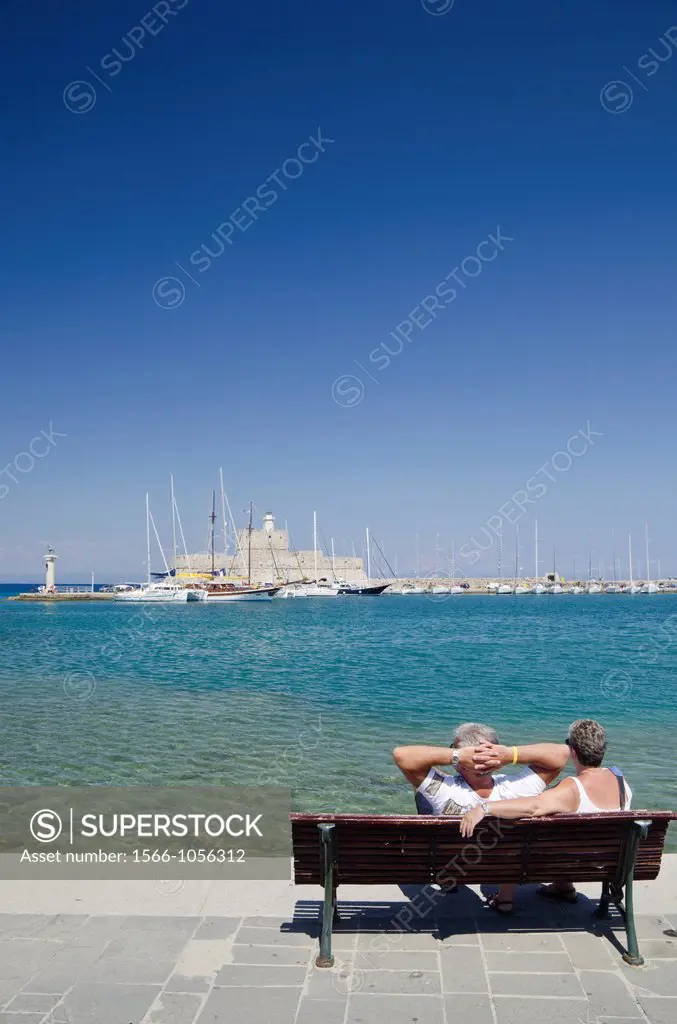 A couple relax on a waterfront bench looking towards the Fort of Saint Nicholas in Mandraki Harbour in Rhodes Town, Rhodes Island, Dodecanese, Greece