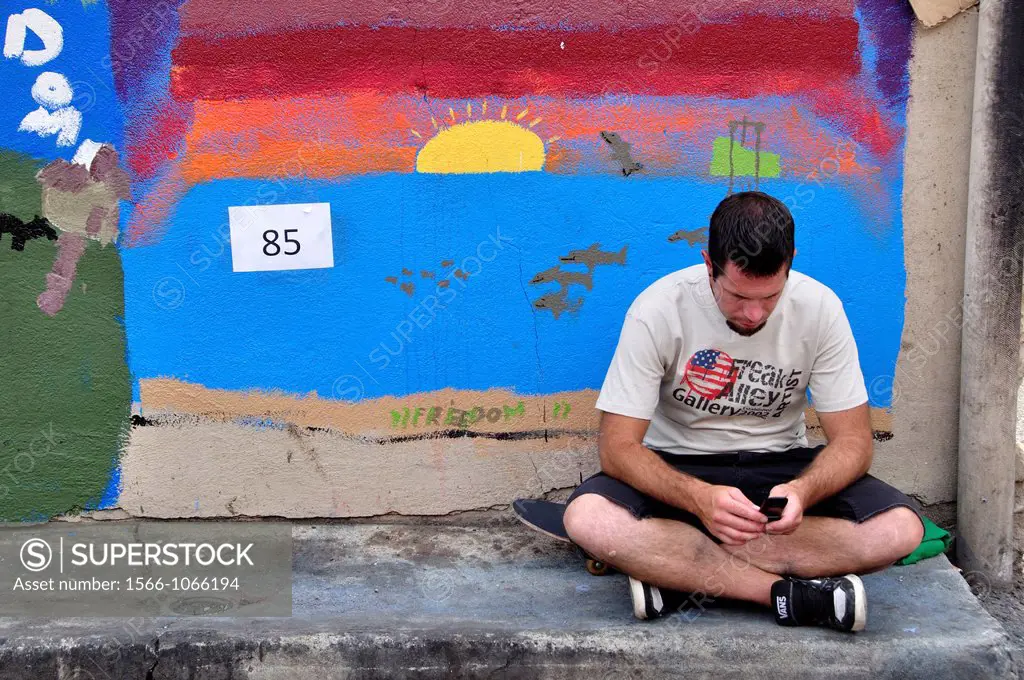 Young man texting a message while sitting next to art on brick wall of building in ´Freak Alley´