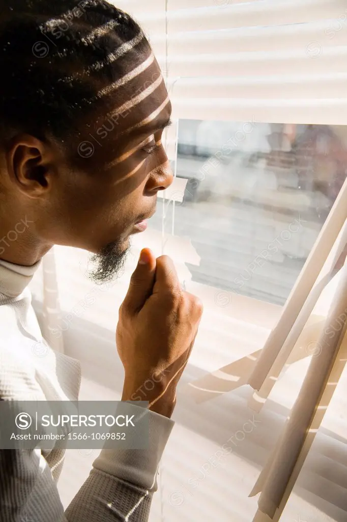 Young black man looking out a window with broken blinds