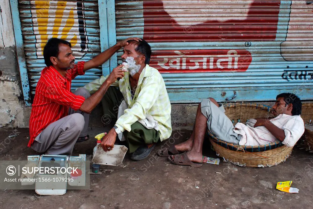 A man is shaving another while a third man is sleeping in a basket. In a street in Bombay, India.