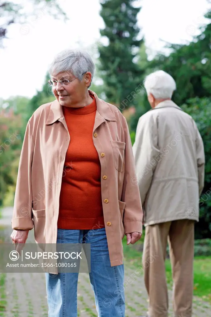 Senior caucasian woman, wearing glasses, looking down with senior man behind her, seen from backside, blurred green background, Hamburg, Germany, Euro...