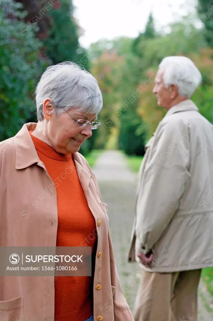 Senior caucasian woman with glasses, looking down with senior man in the background seen from behind, blurred green background, Hamburg, Germany, Euro...
