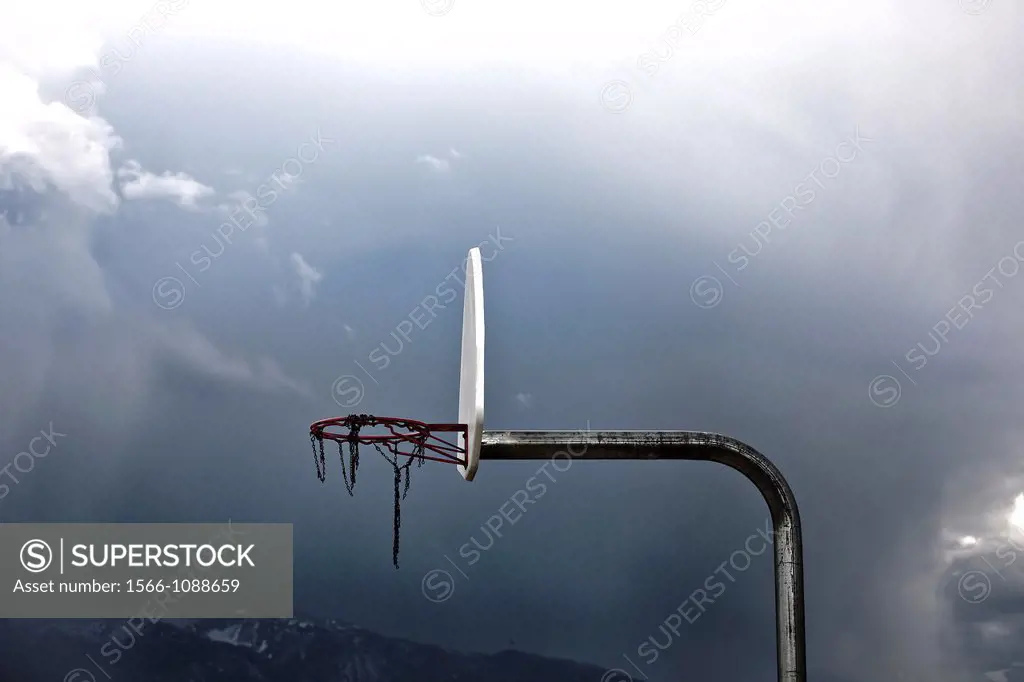 Outdoor basketball hoop with worn down metal net, against a cloudy sky and mountains
