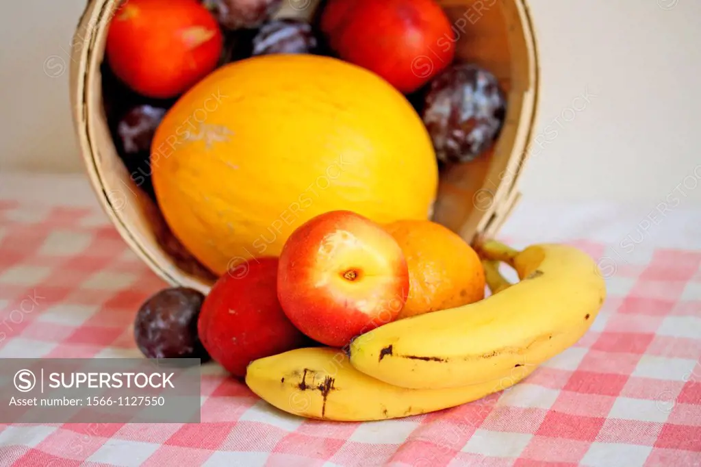 Fruit in a basket  Fruit in basket on its side  Melon surrounded with nectarines and some plums are inside the basket  Bananas and plums lay on the ta...