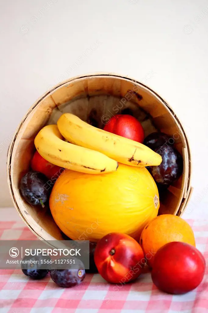 Fruit in a basket that is laid on side for display on red gingham tablecloth  Melon topped with bananas and surrounded with nectarines and plums are i...