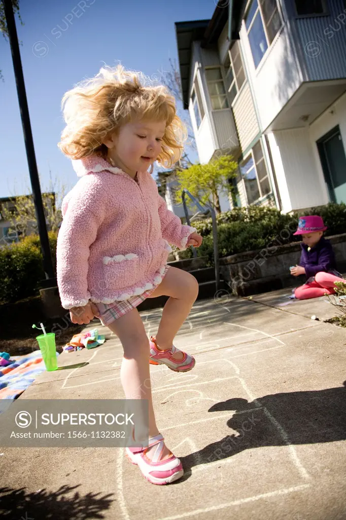 Young girl playing hopscotch infront of her appartment in the sun. Sister playing on the sidewalk behind her.