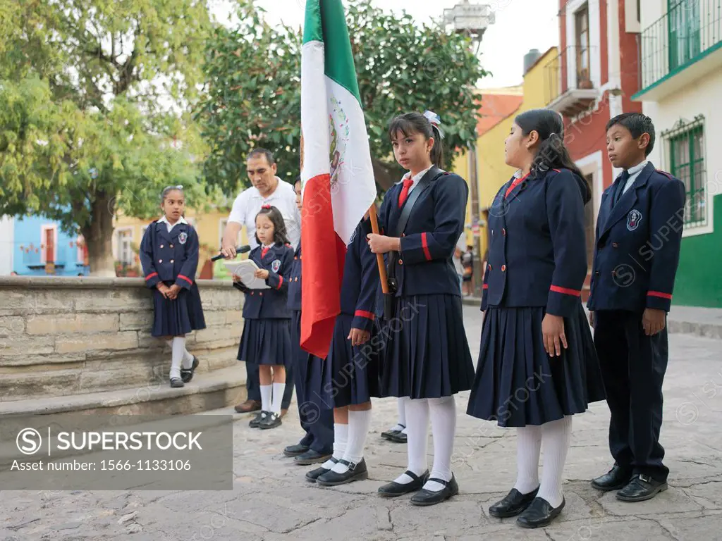 Youg Mexican school children in uniform with a Mexican flag walk around a small plaza in the morning before school begins