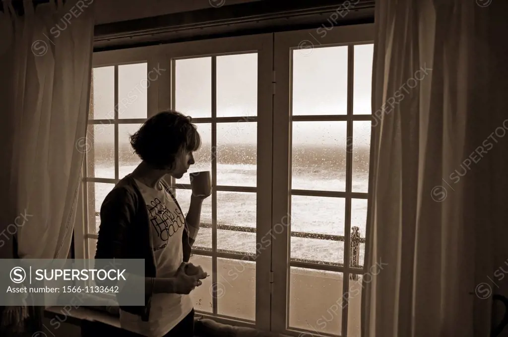 young woman in interior standing in front of completely flooded with water window, Atlantic ocean, beach front hotel room in February, heavy rain and ...