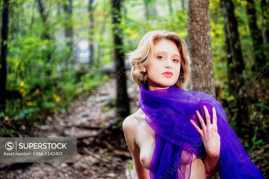 A partially nude 29 year old blond woman wrapped in purple fabric in a forest setting looking at the camera.
