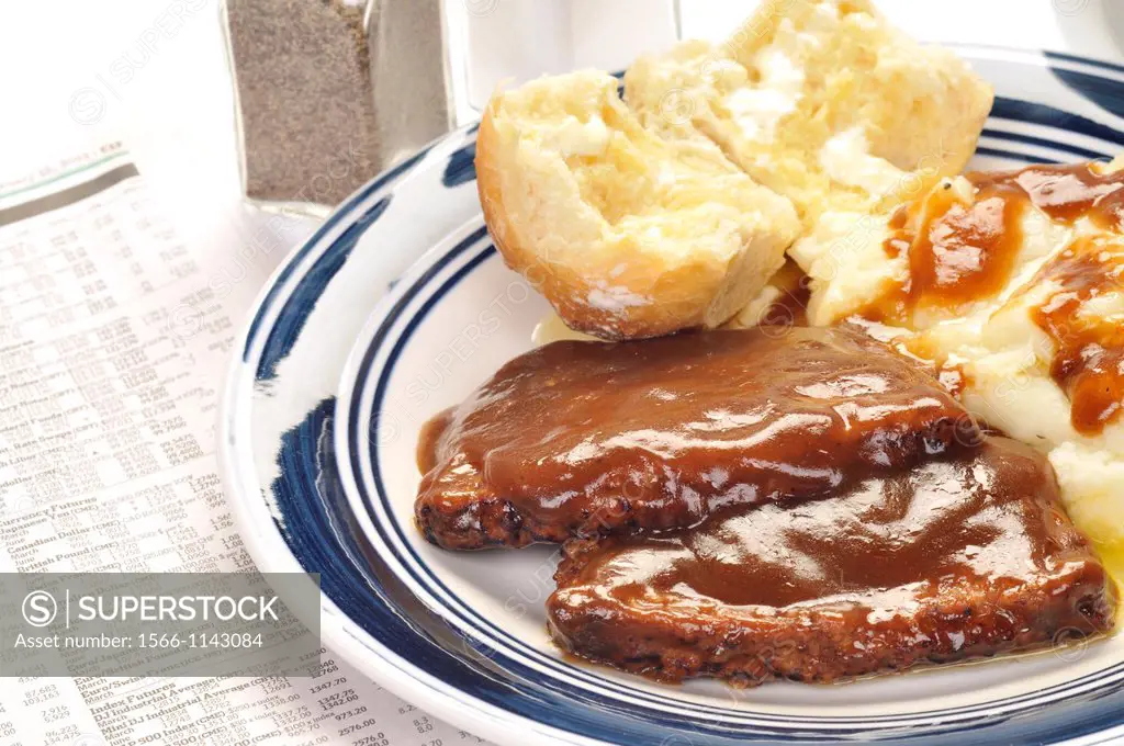 A close up of meatloaf and mashed potatoes topped with gravy and a buttered roll dinner with a cup of coffee and salt and pepper with a newspaper to t...