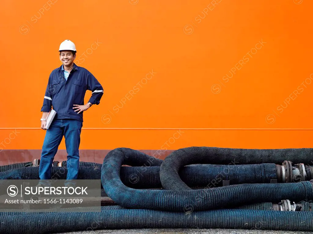 A portrait of a inspector at a commerical container and Palm Oil port in Johor, Malaysia standing on pipes by the hull of a ship