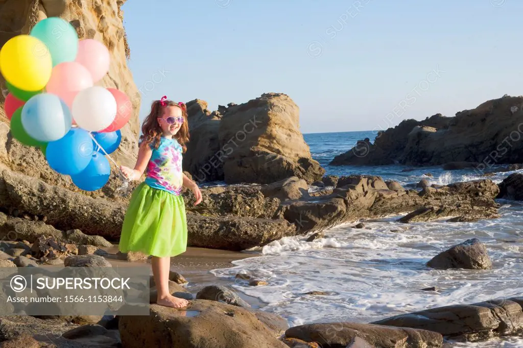 young girl in green skirt standing on rocks at the beach holding balloons