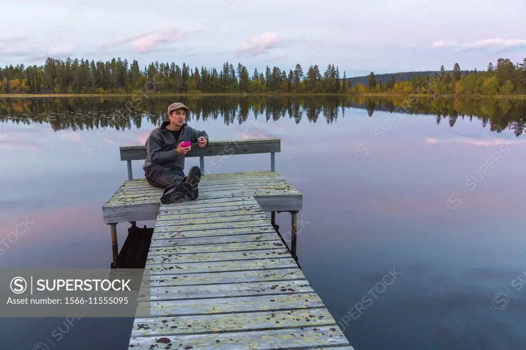 Man sitting on landing stage at a lake in Gällivare, Sweden, drinking coffee.