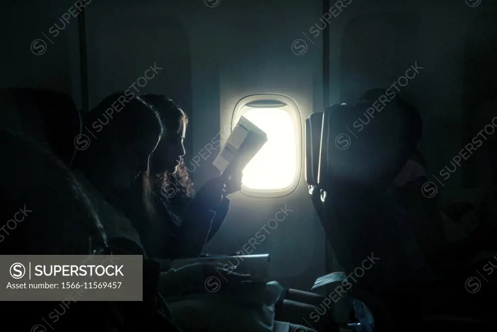 Two young girls sitting in the seats of an airplane reading a backlit of a window.