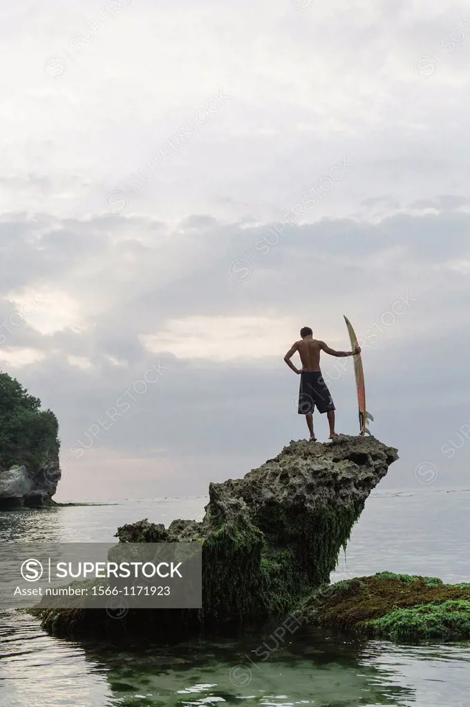 A Balinese surfer perched on top of a rock looking out over the ocean