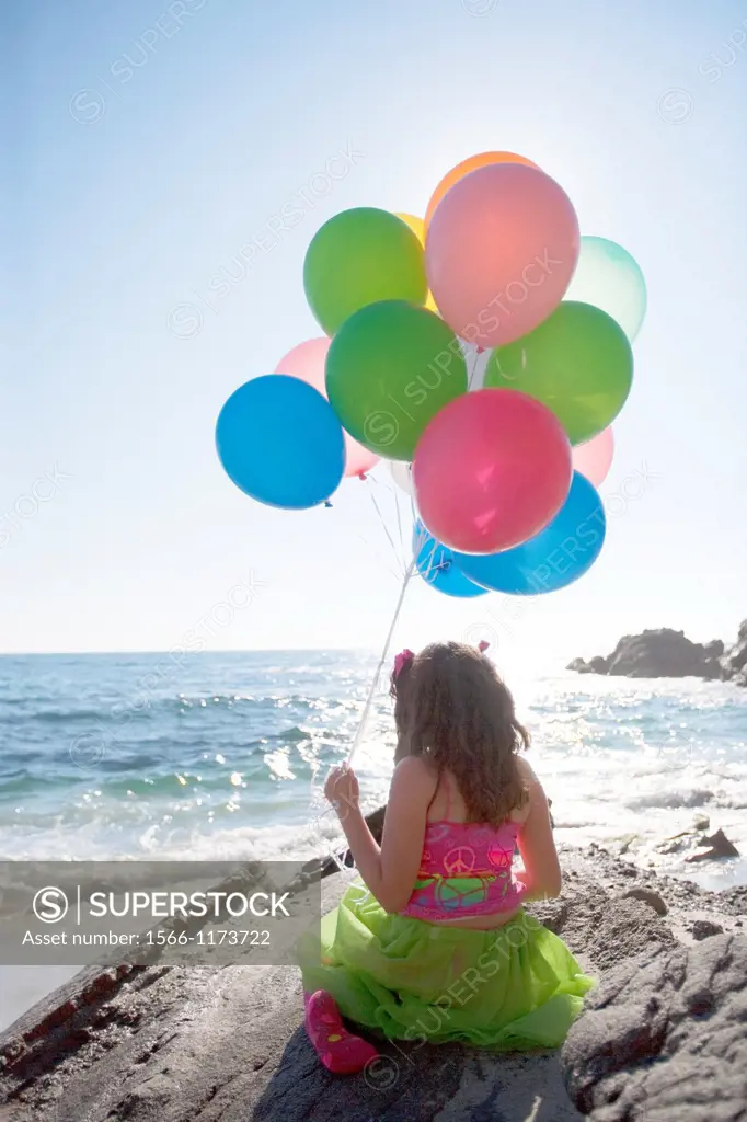 little girl sitting on rocks, holding balloons, overlooking the ocean