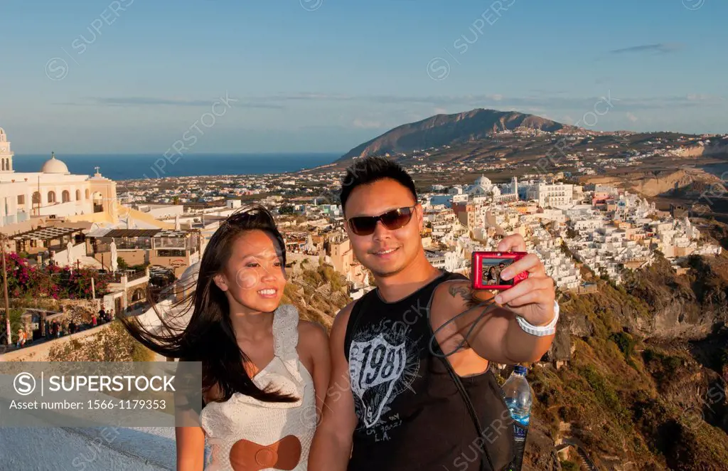 Chinese couple tourists taking picture of themselves overlooking Fira at sunset in Santorini Greece in the Greek Islands