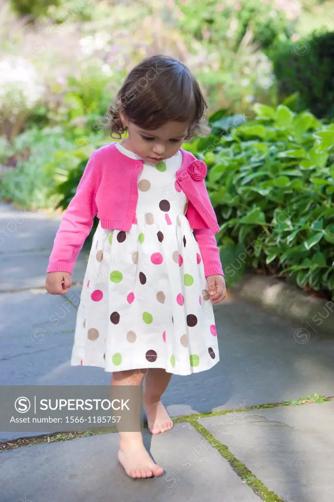 20 Month old girl in poka dots dress with pink cardigan sweater walking bare foot over pavers in garden.