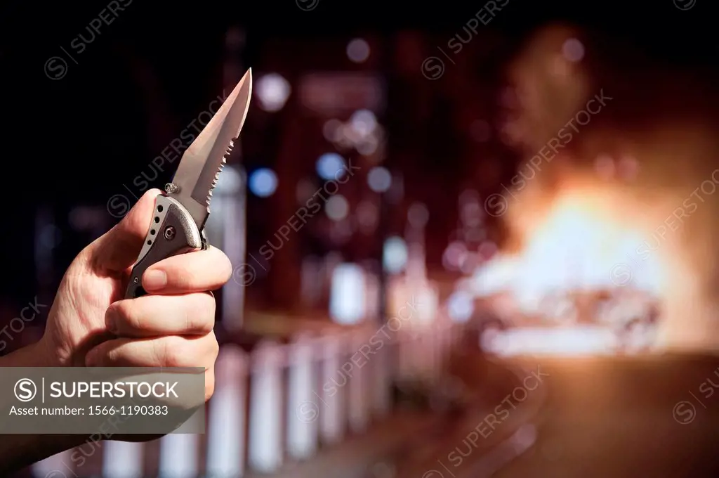 Hand holding a knife with a serrated edge while a car burns in the background  London UK