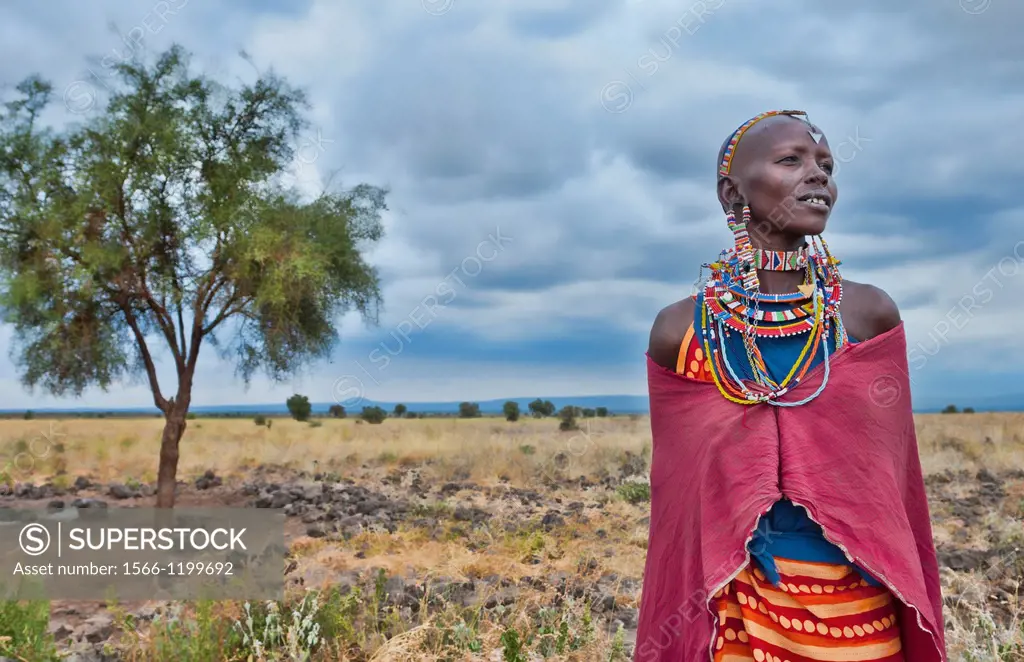 Kenya Africa Amboseli Maasai tribe village Masai woman in red costume dress and beads and tree in remote area of Amboseli National Park safari 1
