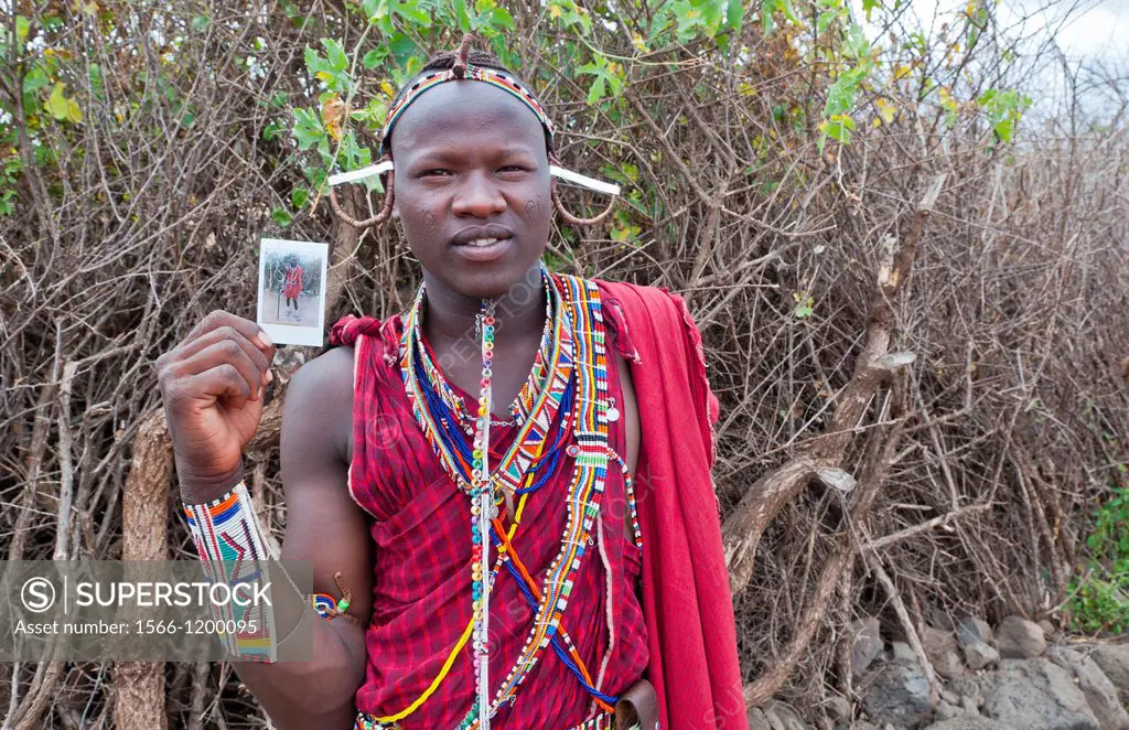 Kenya Africa Amboseli Maasai tribe village Masai man in red costume dress and beads holding Fuji Polaroid in remote area of Amboseli National Park saf...