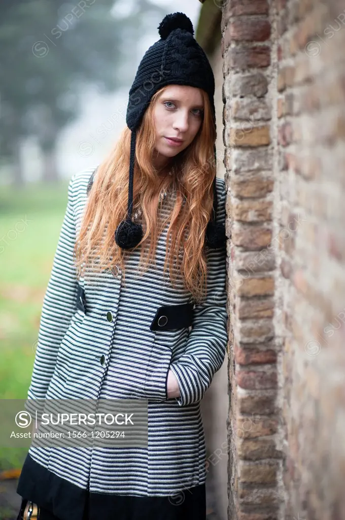 Breda, Netherlands. Young, fashionable and redheaded woman in a city-park, leaning against a wall.