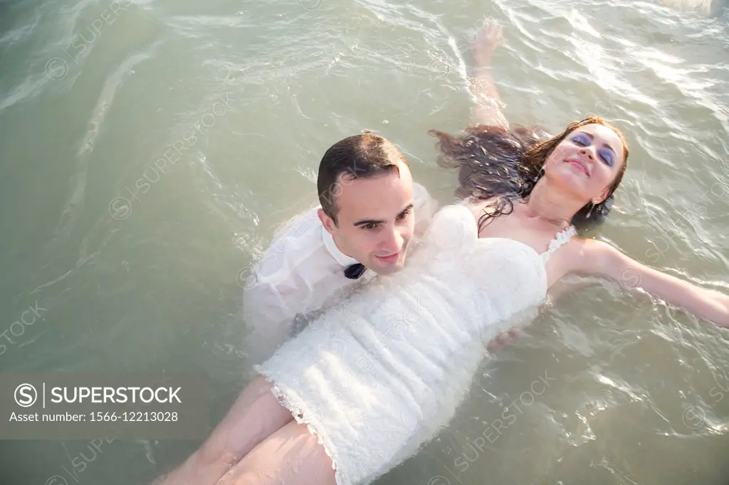 Young man dressed up in a party suit, holding afloat in shalow sea a young woman in a white party dress.