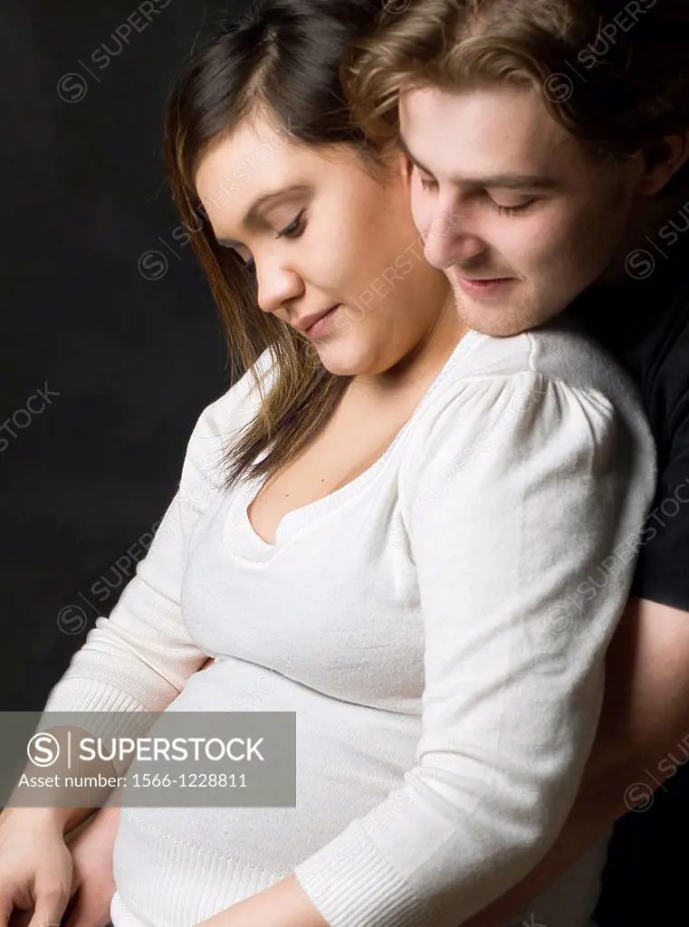 Multicultural couple portrait where the expecting mother is hugged by husband  Both are looking at her belly