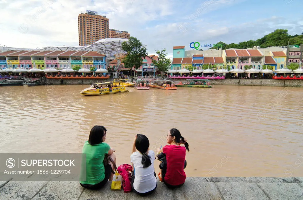 Three women sitting down on the Singapore River bank overlooking the brightly painted entertainment precinct of Clarke Quay, Singapore