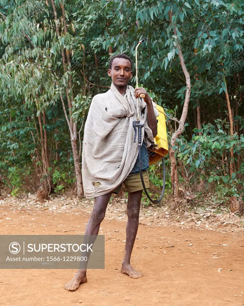 A young barefoot boy carrying a pesticide sprayer and backpack on a dirt road