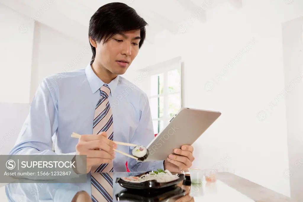 Young Asian businessman eating sushi while communicating on his Tablet computer