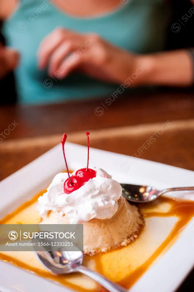 A flan dessert on a plate in a restaurant win a woman´s hands in the background.