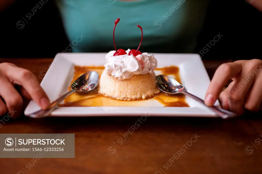 A flan dessert on a plate in a restaurant win a woman´s hands next to the plate.
