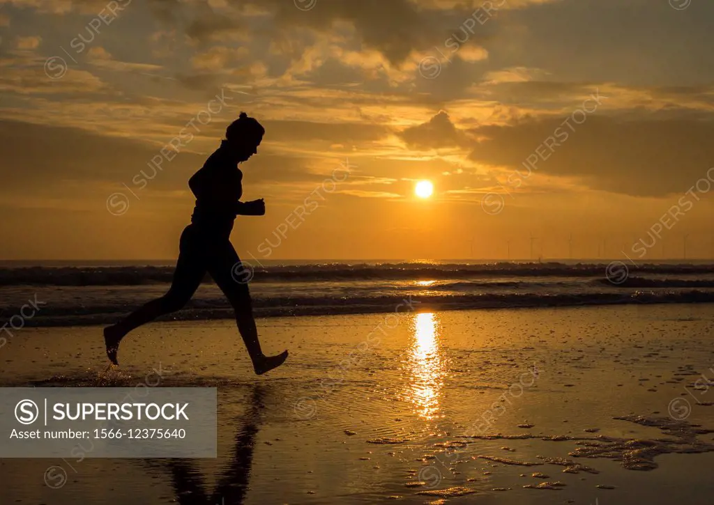 Woman running barefoot along shoreline at sunrise in the North Sea at Seaton Carew, North east England, United Kingdom.