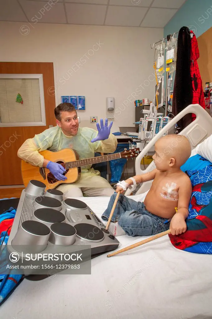 Wearing a sterile gown and rubber gloves, a music therapist playing a guitar entertains a young boy chemotherapy cancer patient at CHOC Children´s Hos...