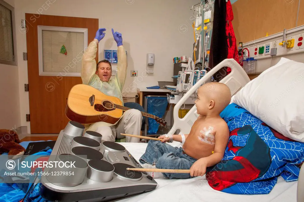 Wearing a sterile gown and rubber gloves, a music therapist playing a guitar entertains a young boy chemotherapy cancer patient at CHOC Children´s Hos...