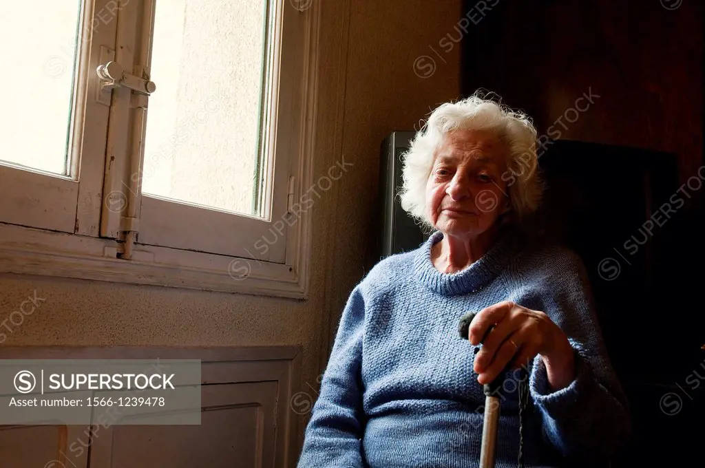 Portrait of old woman at home, looking at the camera, sitting by the window.