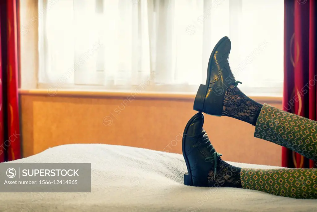 Closeup of feet of woman with oxford shoes lying on a bed in a relaxing position and a window in the background.