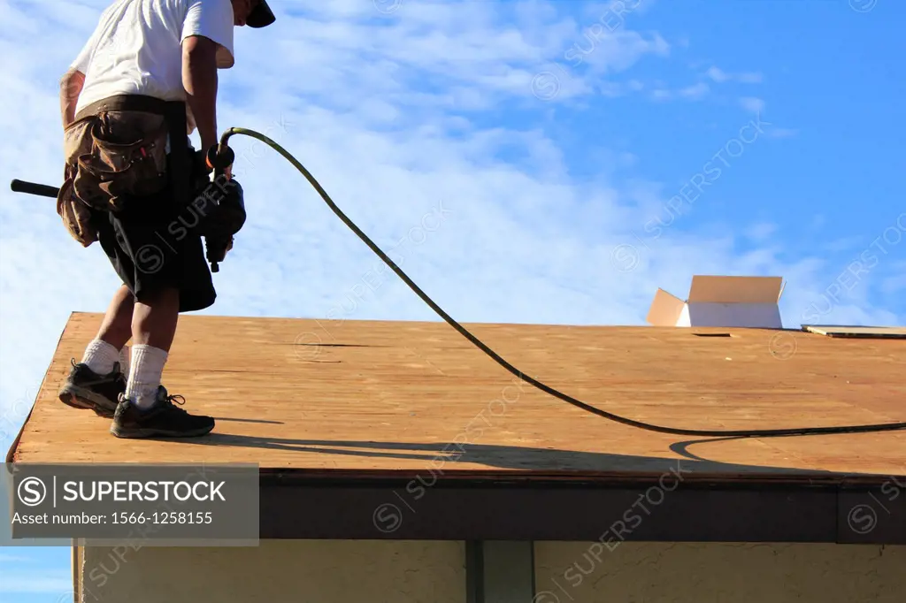 a man or roofer using a nail gun and working to replace the plywood or wood or covering and shingles on a house roof