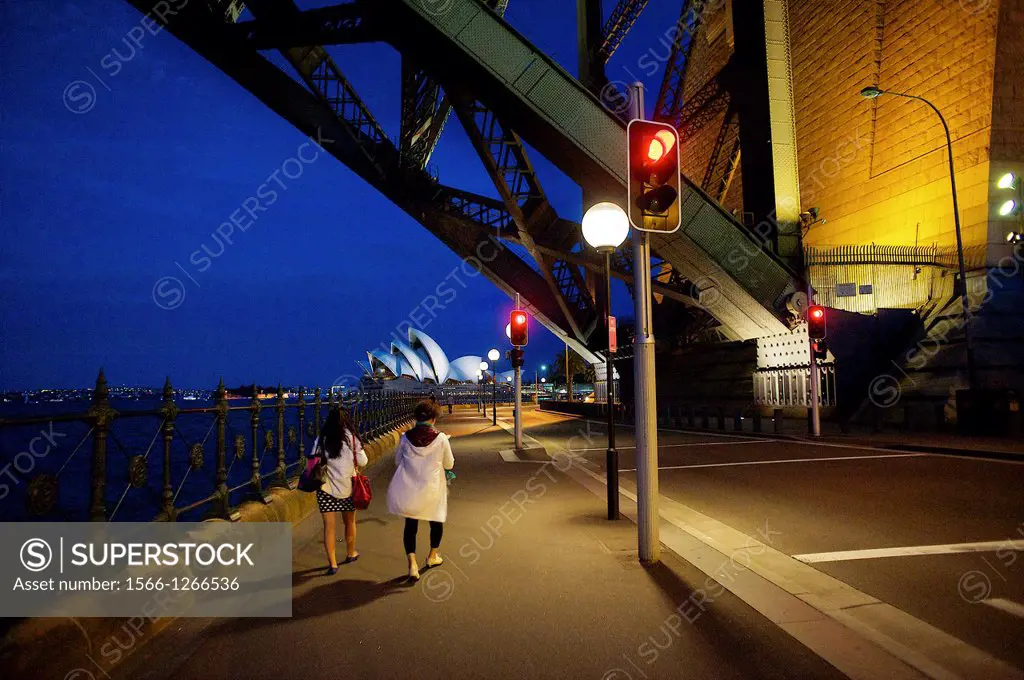 twilight view of two women walking below the Sydney Harbour Bridge with the illuminated Opera House in the distance