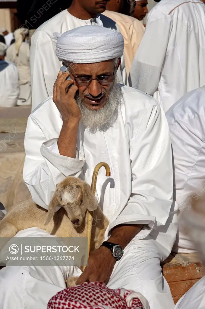 Omani man with a goatling in his arms making a telephone call with a cellular phone at the goat market, Nizwa, Sultanate of Oman